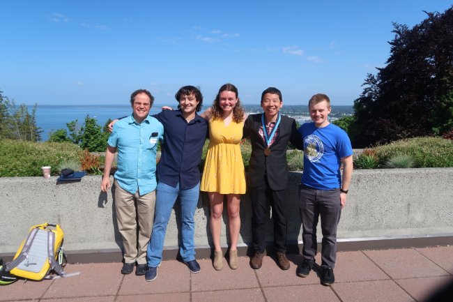 A group of five people is posing for a photo on a rooftop. The men are wearing casual clothes, while the woman is dressed more formally. The people are all smiling. In the background, there is a view of Bellingham and Bellingham Bay, looking north.
