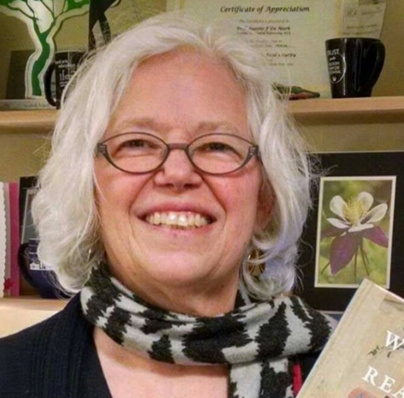  The photo shows a woman with gray hair and glasses smiling. She is wearing a black shirt with a gray and black striped scarf. There are books and awards on the shelf behind her.