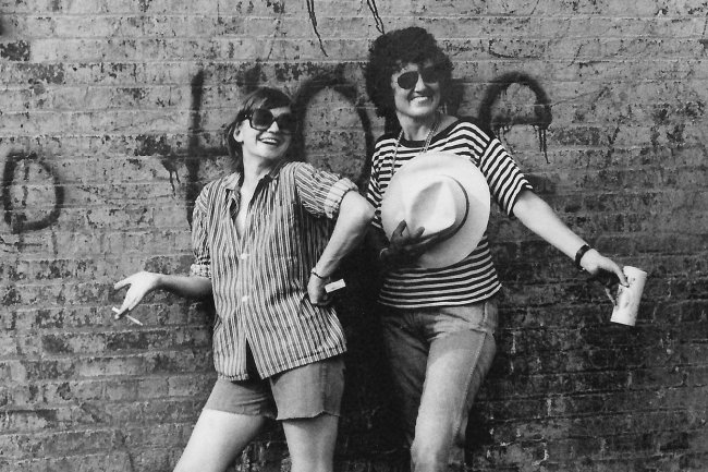 Two smiling women pose in front of a brick wall in New York City in the 1970s.