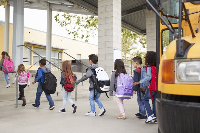 Seven elementary school-aged children wearing backpacks and exiting a school bus on their way into school.