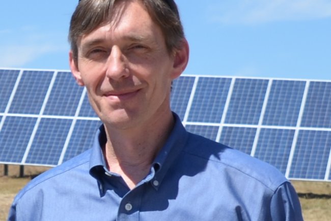 Darrin Magee smiles at the camera; behind him in the sun is a row of solar panels