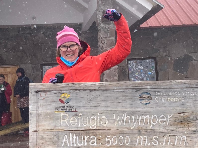 Amy Carbajal lifts her arm in celebration as she reaches the second refugio on Chimborazo, with sleet falling and a structure in the background.