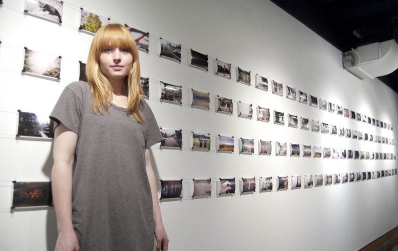 AS Productions VU Gallery Coordinator Ashley Hollender poses in front of a wall of photos for the "An Experiment" exhibit opening in the VU Gallery Monday, January 9. The exhibit features photos taken by random people who were handed cameras and given fre