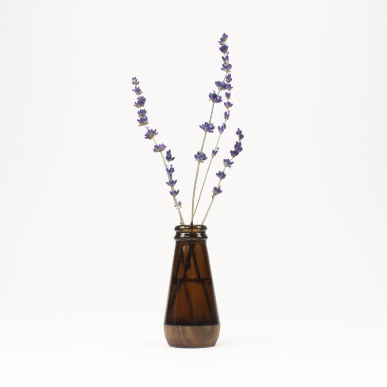 A small brown glass bud vase holds several stems of lavender. The lavender has purple flowers and green stems and leaves. The vase is made of recycled glass and has a wooden base. The vase is sitting on a white table or surface. The background is white.
