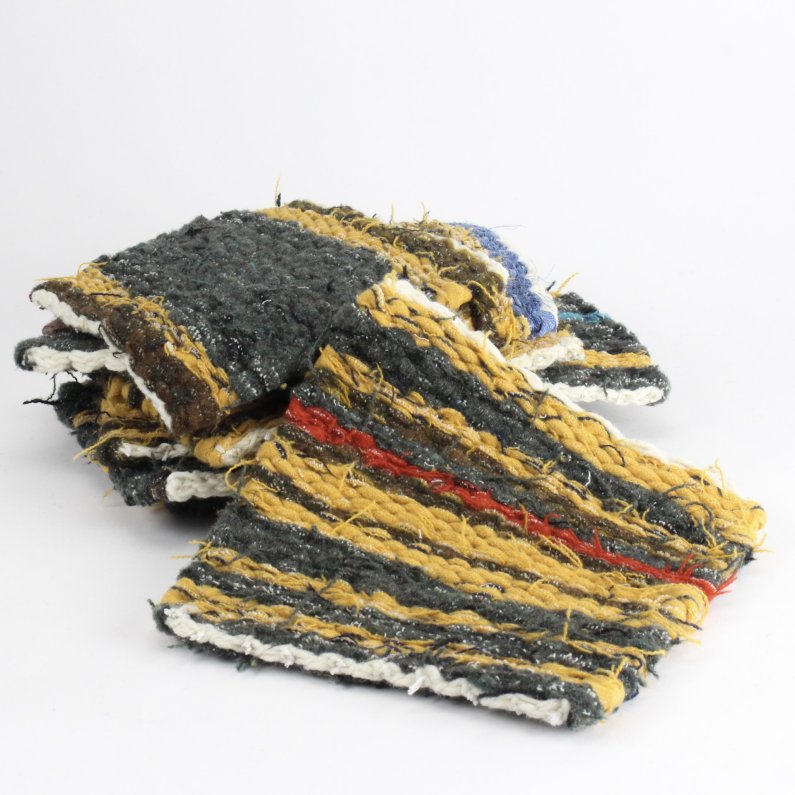 The image shows a handwoven rag rug made of recycled materials. The rug has a striped pattern in brown, yellow, blue, and red. The edges of the rug are frayed and uneven. The rug is lying on a white background.