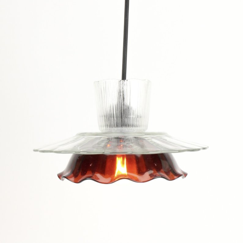 The image shows a handmade glass pendant lamp. It consists of two glass shades - a clear ribbed glass shade and a brown ruffled glass shade. The shades are attached to each other with a metal ring. The lamp is suspended from a black cord. The light bulb is located inside the brown glass shade and is turned on, casting a warm glow.