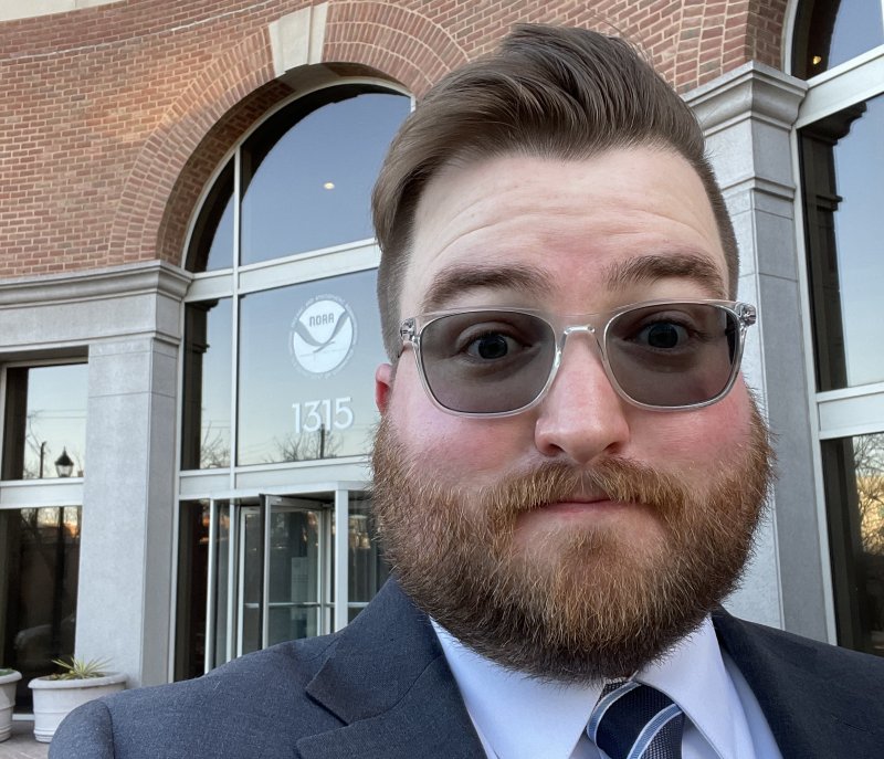 Michell Hebner, wearing a suit and tie and first-day-of-a-new-job haircut, takes a selfie in front of a building with a NOAA logo