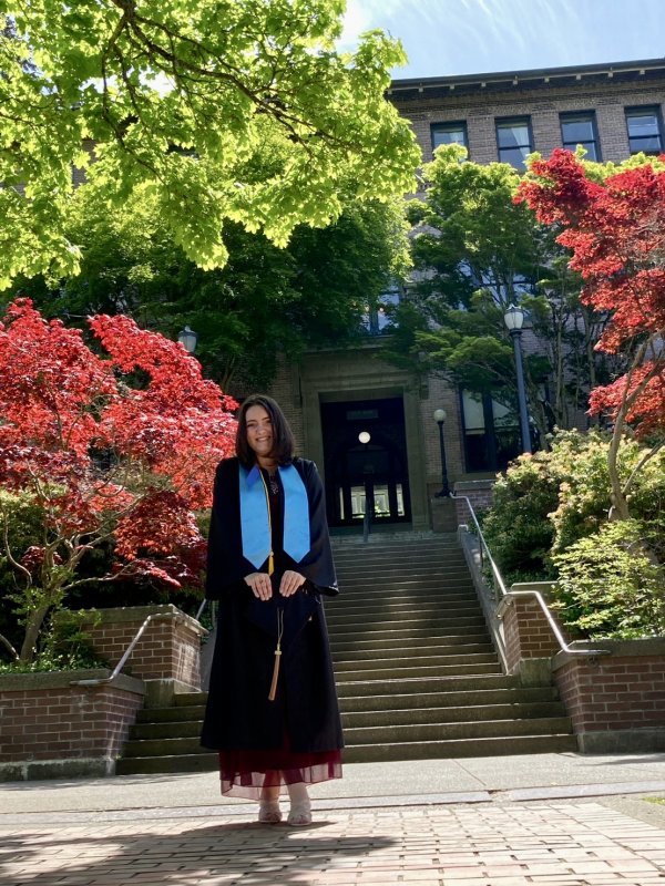 Alina Sokolova smiles for the camera, wearing her graduation regalia and standing in front of the Old Main steps. 