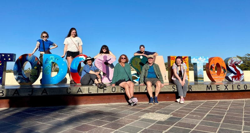 The study abroad group sits in the sun in front of a colorful Todos Santos sign.