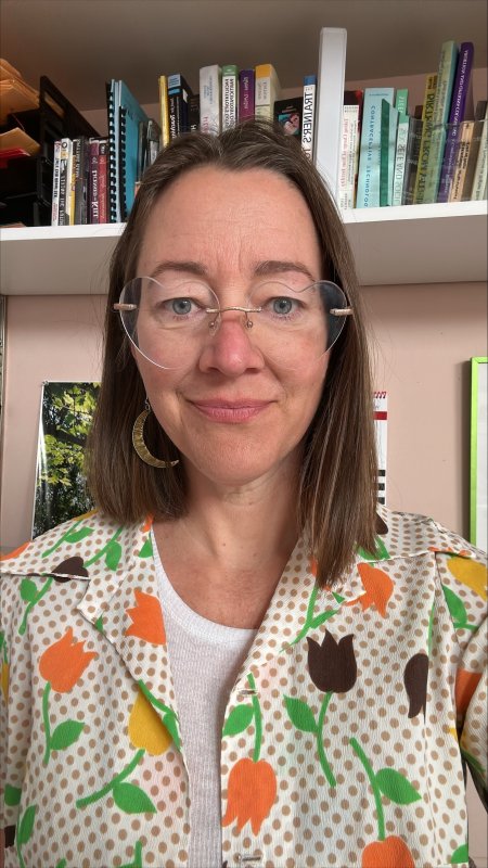A woman with brown hair and glasses is smiling at the camera. She is wearing a white shirt with a colorful floral pattern and a crescent moon earring. There are bookshelves and papers in the background.
