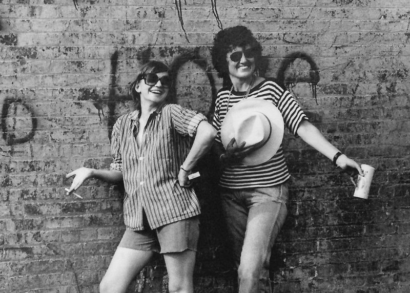 Two smiling women pose in front of a brick wall in New York City in the 1970s.