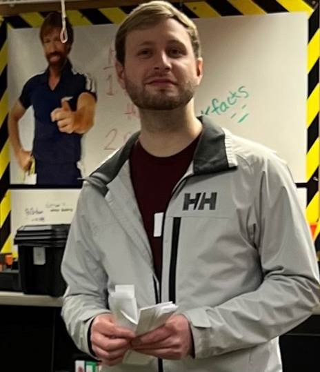 The image shows a man standing in front of a white board. He is wearing a gray Helly Hansen jacket, a maroon shirt, and has short brown hair. He has a beard and his eyes are looking at the camera. There is a poster of Chuck Norris behind him on the wall.