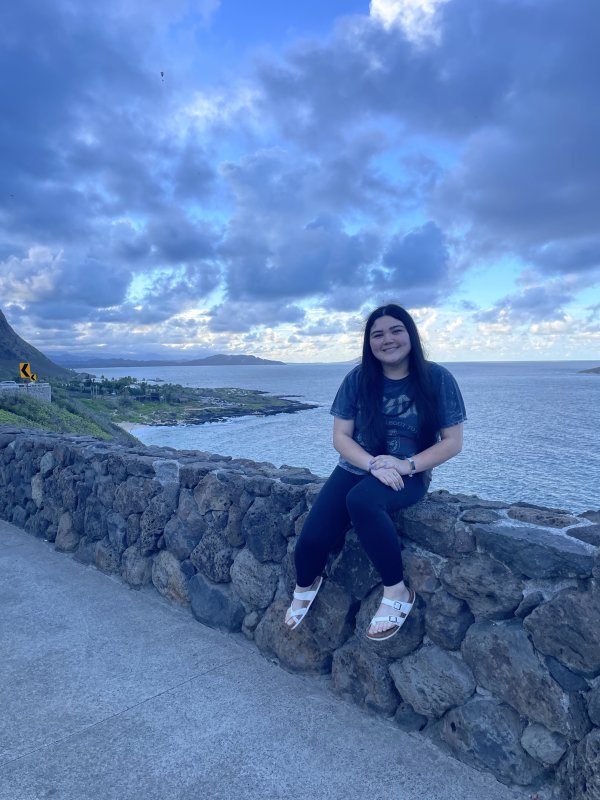 Mehana Tamashiro sits on a waterfront ledge, smiling for the camera 