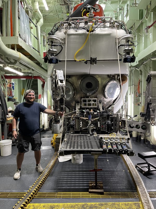 Mitch Hebner stands smiling next to the Alvin submarine, loaded into the interior of a ship