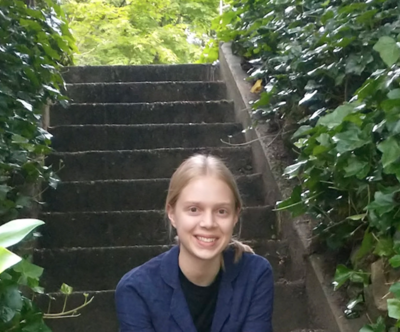 Petra Ellerby smiles for the camera while sitting on concrete steps surrounded by leafy green ivy