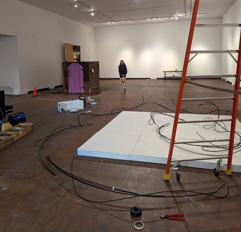 A student on a razor scooter rolls through an empty-walled Western Gallery. The floor is cluttered with artwork, cables and ladders. 