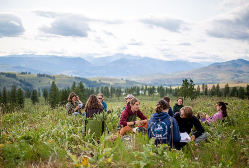 Students sit in a ring in a grassy meadow; behind them are the green foothills of the Methow Valley.