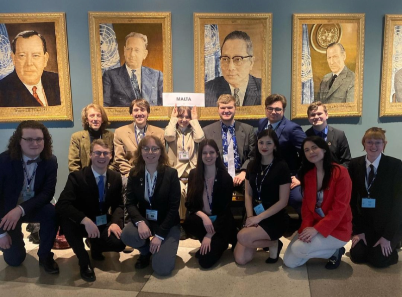 A group of students pose for a photo in front of a wall of portraits of former UN Secretary-Generals. The students are all wearing suits or formal dresses. In the center of the group, one student is holding a sign that says "Malta."
