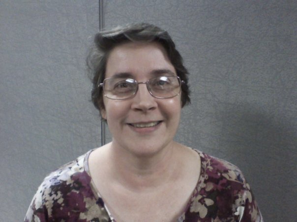 The photo shows Valerie Klein, who has short gray hair and glasses. She is wearing a purple shirt with a floral pattern. The woman is smiling and looking at the camera. The background is a gray wall.