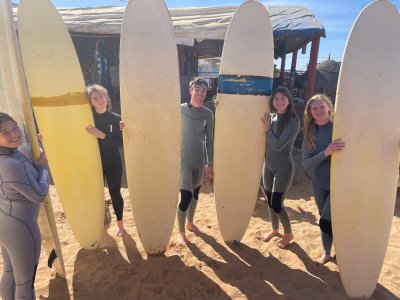 Four students in wet suits stand on a sunny beach next to surfboards 