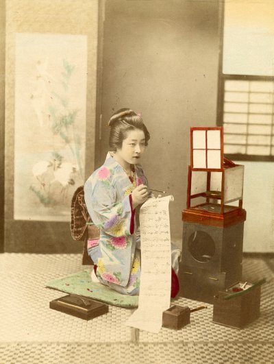 A Japanese woman holds a scroll of caligraphy while she kneels on a reed mat.