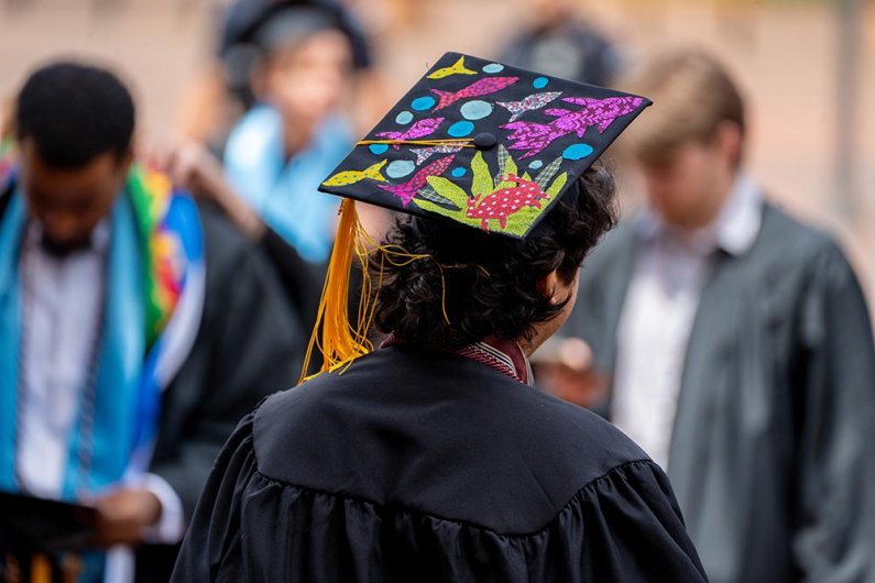 A graduate student wearing a black graduation cap decorated with colorful fish and sea life.