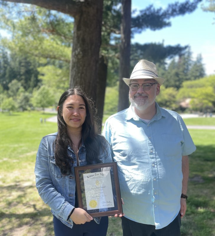 A woman and a man are standing in a park. The woman is holding a framed certificate. They are both smiling.