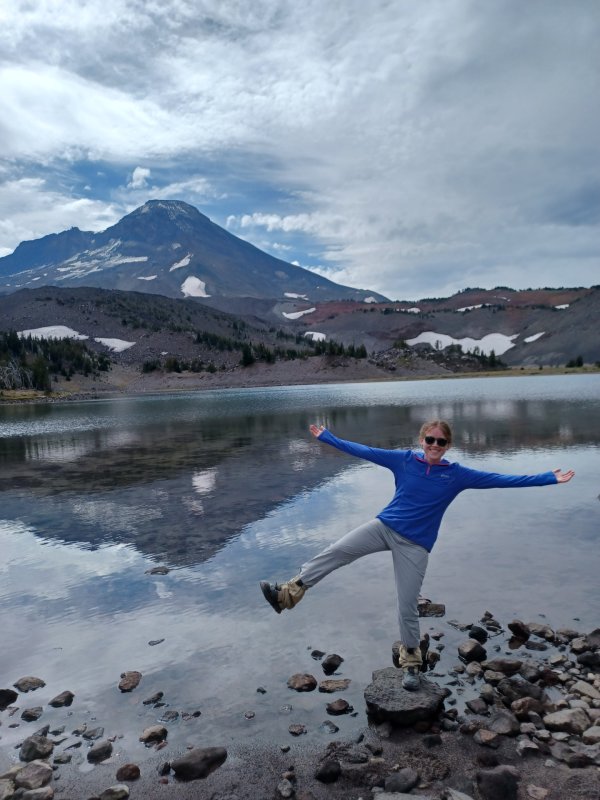  A woman standing on the edge of a lake with a large snow-capped mountain in the distance. She is wearing a blue shirt and gray pants and has her arms outstretched.