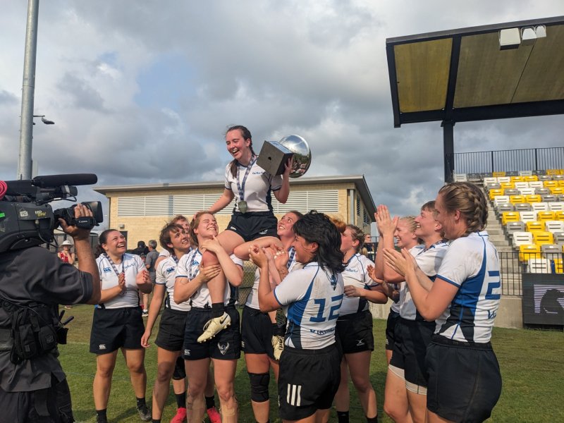 A group of female rugby players are celebrating their victory. They are hoisting one of their teammates up on their shoulders while others surround her, holding the trophy. The players are all wearing white and blue uniforms. They are standing on a field that is surrounded by stands. A camera operator is filming the celebration.