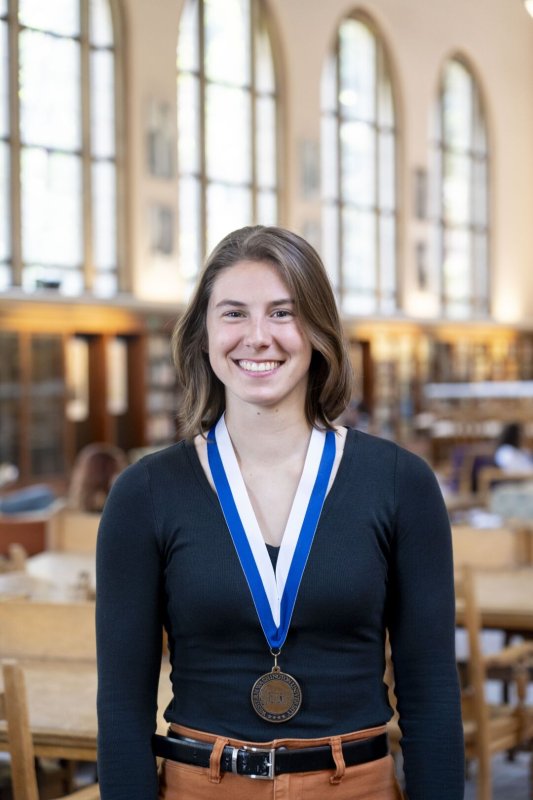 A young woman smiles while wearing a medal and stands in a library.