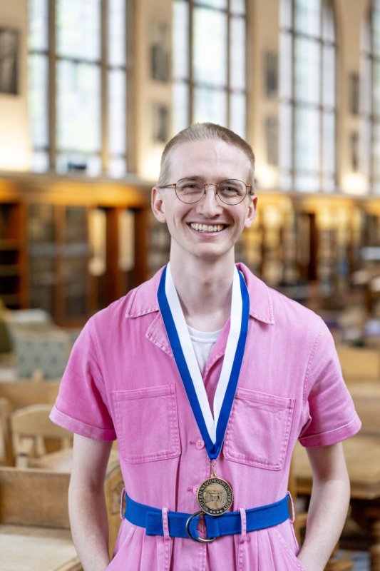 A young man in a pink jumpsuit and glasses smiles while wearing a medal. Behind him are the walls and shelves of a library.