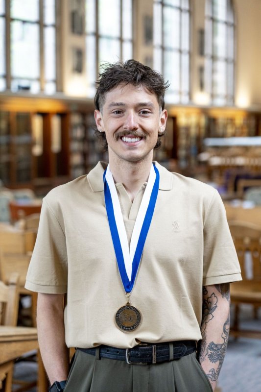 A young man is smiling and wearing a medal. He is standing in a library and there are bookshelves and a large window behind him.