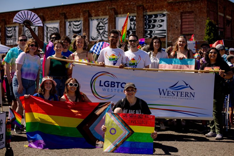 A group of people are posing for a photo while holding a banner that says “LGBTQ+ Western” and a rainbow flag. The group is standing in front of a brick building.