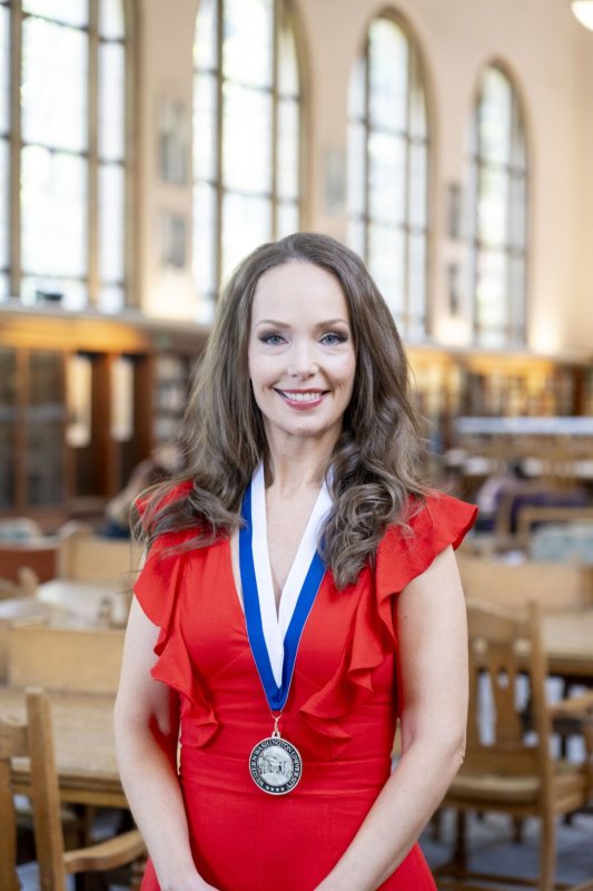A smiling woman with long, wavy brown hair is wearing a red dress and a blue and white ribbon with a medal on it. She is standing in a library with many empty wooden tables and chairs.