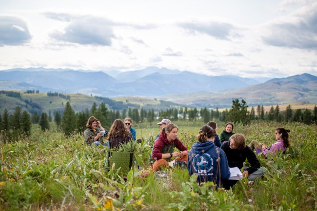 Students sit in a ring in a grassy meadow; behind them are the green foothills of the Methow Valley.