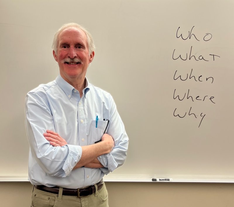 George Erb poses in front of a white board on campus; written on the white board are the words "who, what, when, where, why."