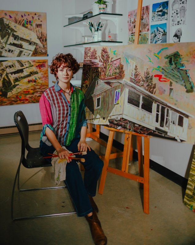 Tesla Kawakami, paintbrush in hand, sits in her studio surrounded by paintings