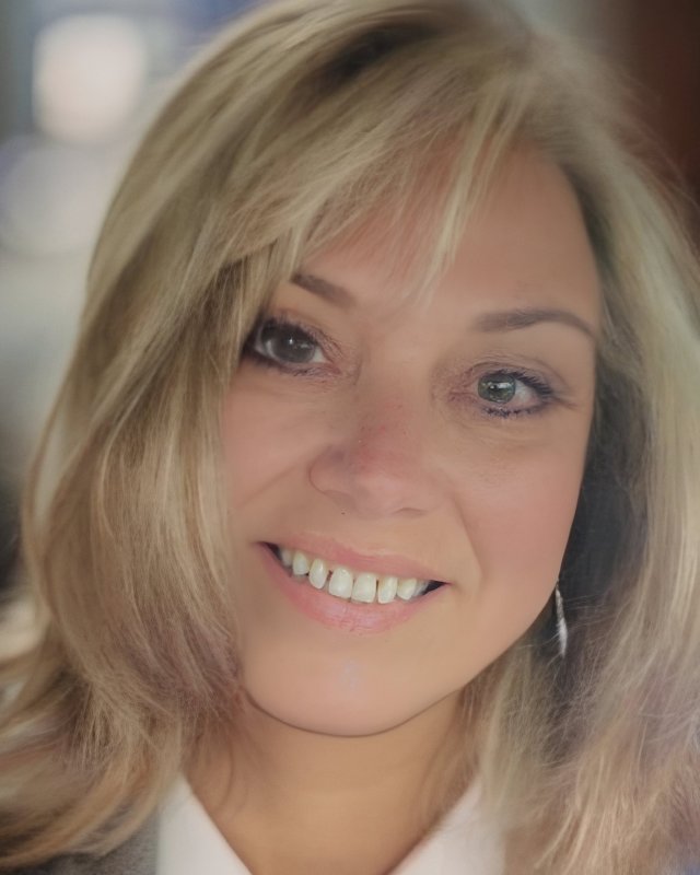 The image shows Traci Rogstad, a woman with blond hair and green eyes. She is smiling and looking at the camera. She is wearing a white shirt. The background is blurred.