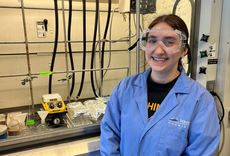 Aimee Long, wearing a lab coat and goggles, smiles at the camera in a chemistry lab