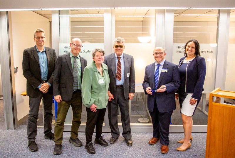Group photo taken moments before the ceremonial ribbon cutting with people standing in front of the blue ribbon tied between the glass consultation rooms of the new Hub. From left to right: Brad Johnson, David Patrick, Kathy Kitto, Dennis Waller, John Danneker, and Kim O’Neill